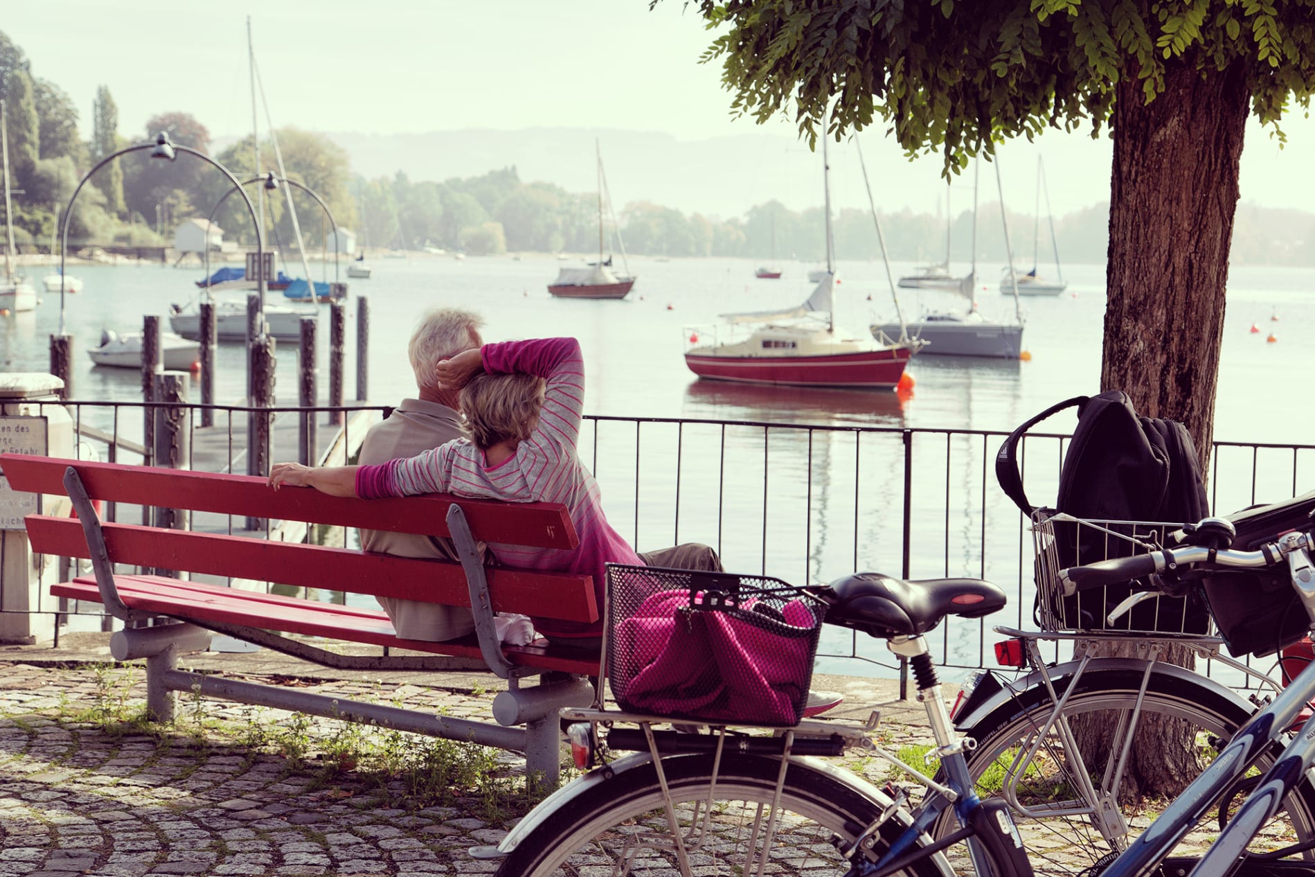 couple sitting on a bench overlooking a serene lake with boats, bikes parked nearby amidst a peaceful, natural setting
