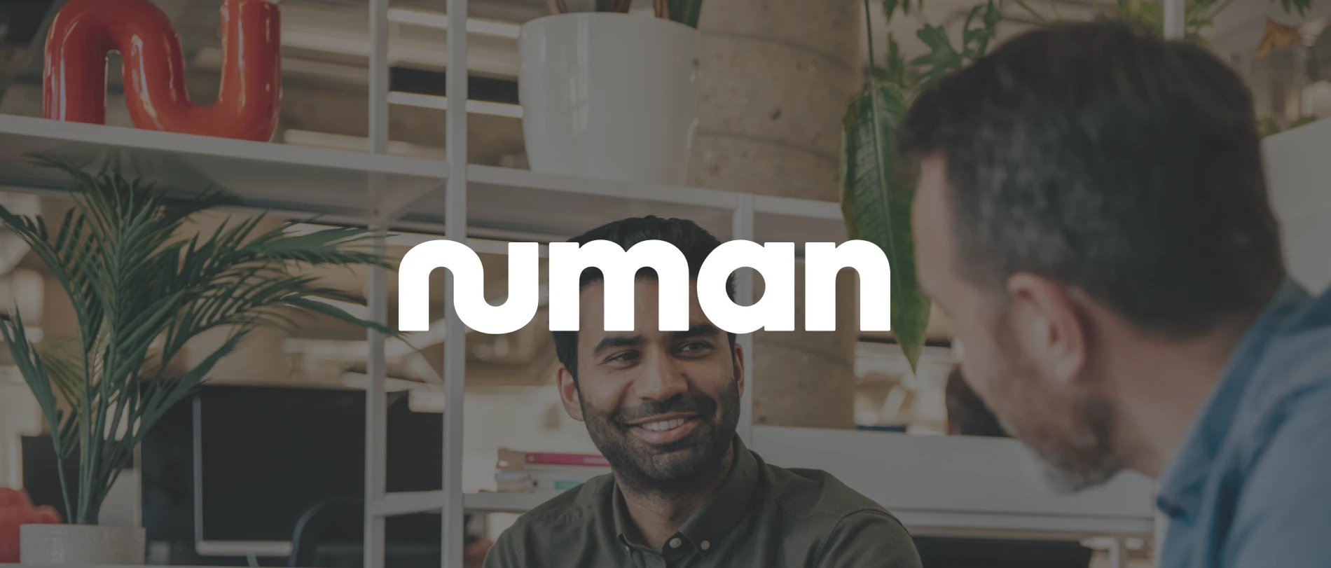 numan logo displayed in a modern office setting with two men engaging in conversation by a plant, interactive office environment with greenery