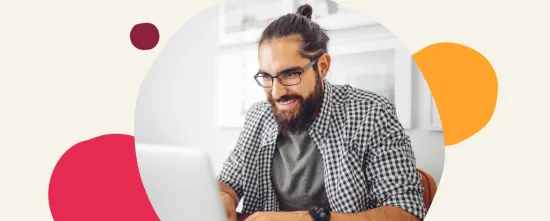 man with glasses smiling while working on a laptop in a home office setting, casual work environment with artistic design elements