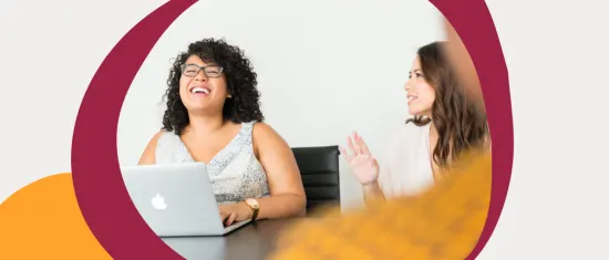 smiling women in a meeting, one with glasses, laptop on table, friendly interaction, modern office setting, happy coworkers collaborating in office