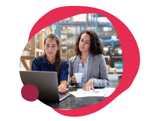 working women discussing data analysis on laptop in warehouse, one holding a coffee cup, business meeting with charts and graphs