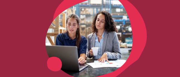 business women collaborating, analyzing data on laptop with documents in a warehouse setting
