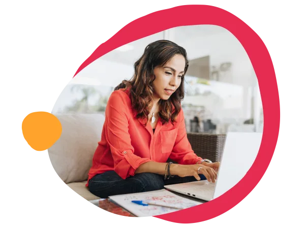 working woman in red shirt using laptop, focused expression, casual setting with notebook and pen on table, remote work lifestyle