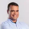 smiling man in a light blue shirt, with short black hair against a neutral background, friendly expression, portrait format