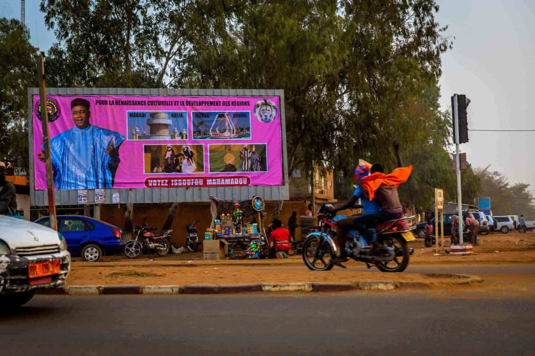 Twee aanhangers van Hama Amadou rijden voorbij een billboard van huidig president Issoufou Mahamadou, dat culturele renaissance en ontwikkeling van de regio’s belooft.