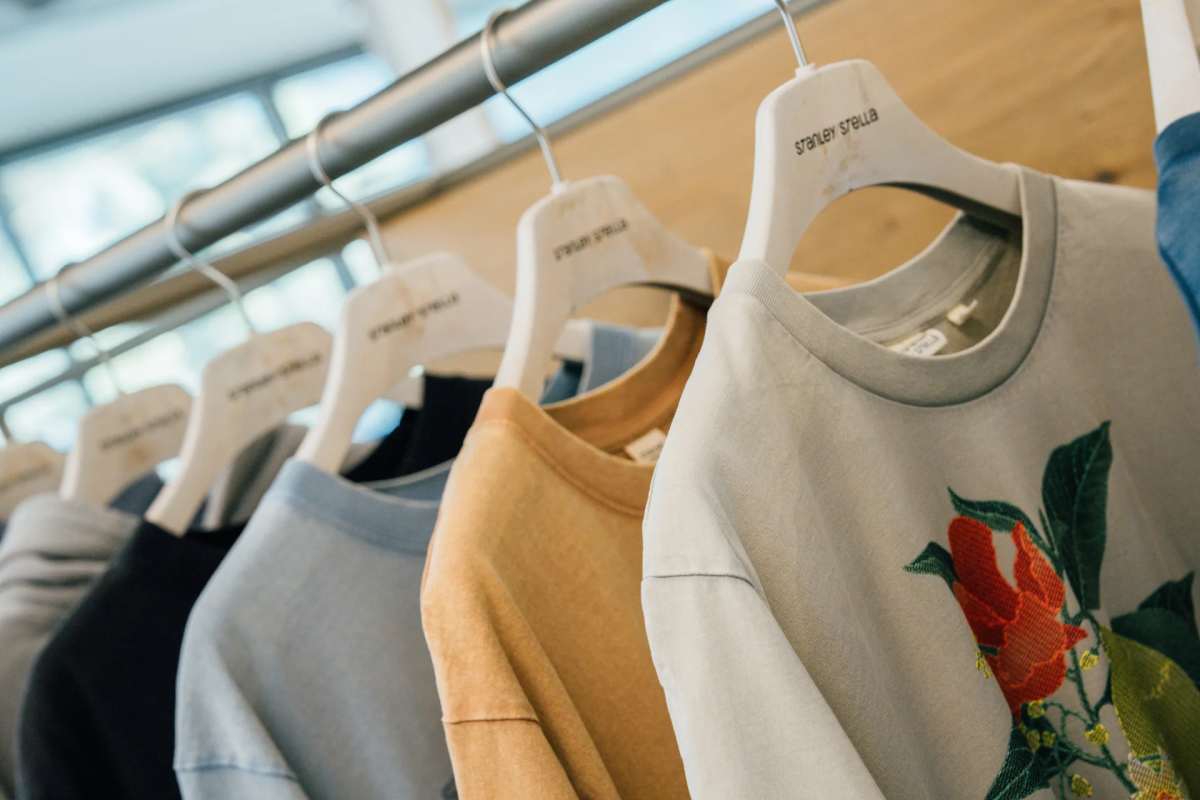 A clothing rack holding multiple Stanley Stella branded sweatshirts in various colors including light gray with floral embroidery, mustard yellow, light blue, navy, and white, displayed on white branded hangers in a bright retail or showroom setting.