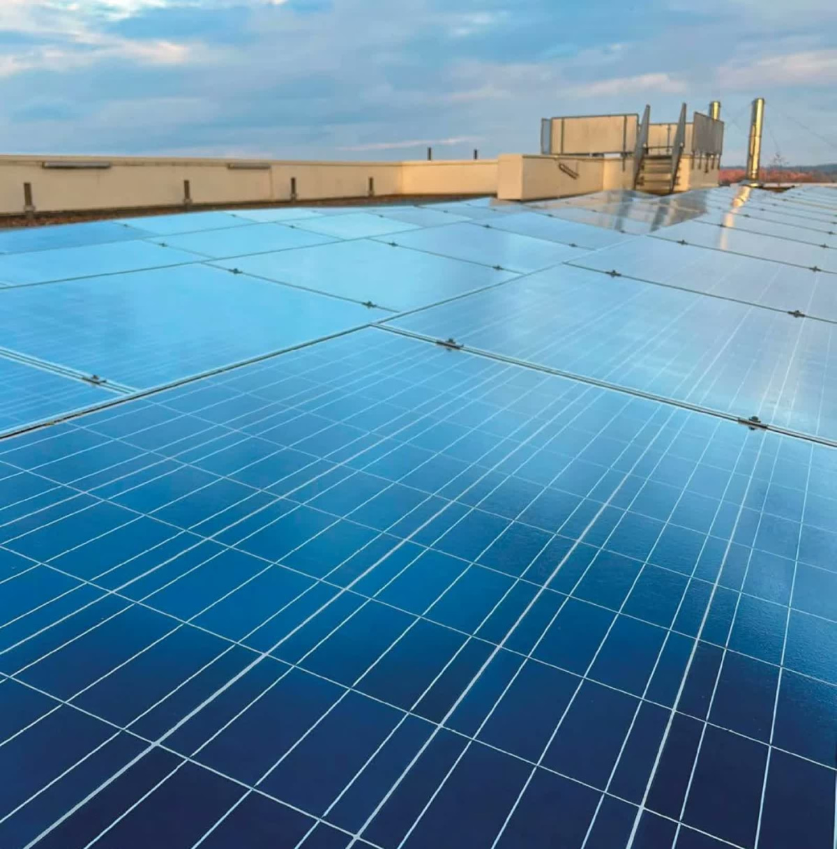 A large array of blue photovoltaic solar panels covering a flat commercial rooftop, with HVAC equipment and a stairwell structure visible in the background against a partly cloudy sky.
