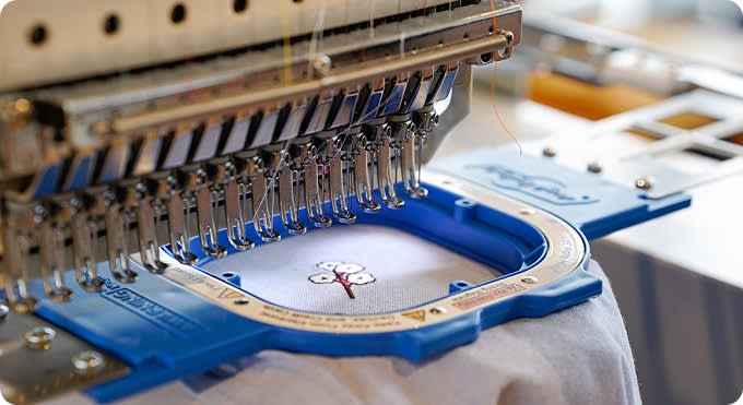 A multi-needle industrial embroidery machine with a blue hoop frame stitching a small floral or cotton-boll motif onto white fabric.