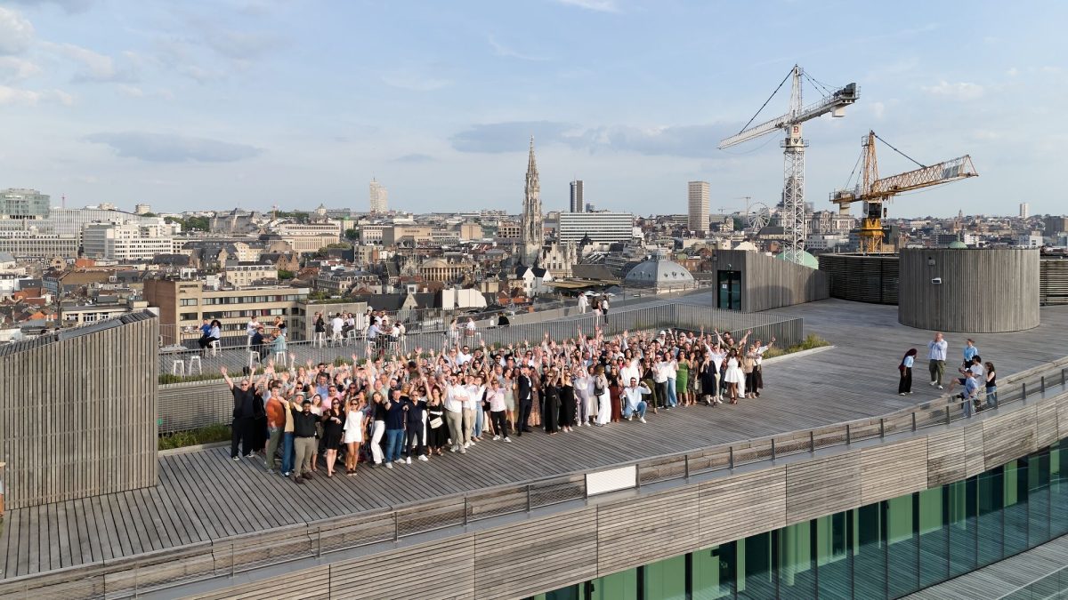 Een grote groep van ongeveer honderd mensen op een dakterras met houten vlonder, zwaaiend naar de camera, met op de achtergrond een Europese skyline.