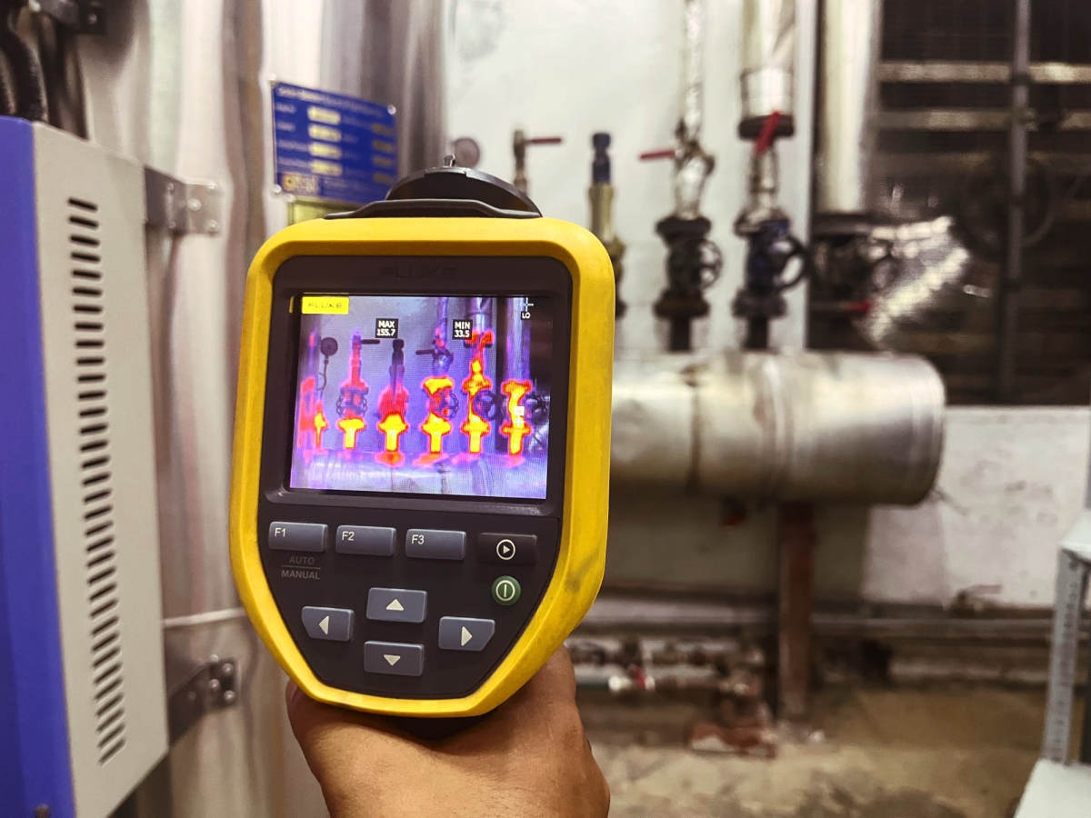A hand holds a yellow Fluke thermal imaging camera displaying a false-color infrared heat map of industrial pipes and valves, with the same pipes and valves visible in the background of a utility room.