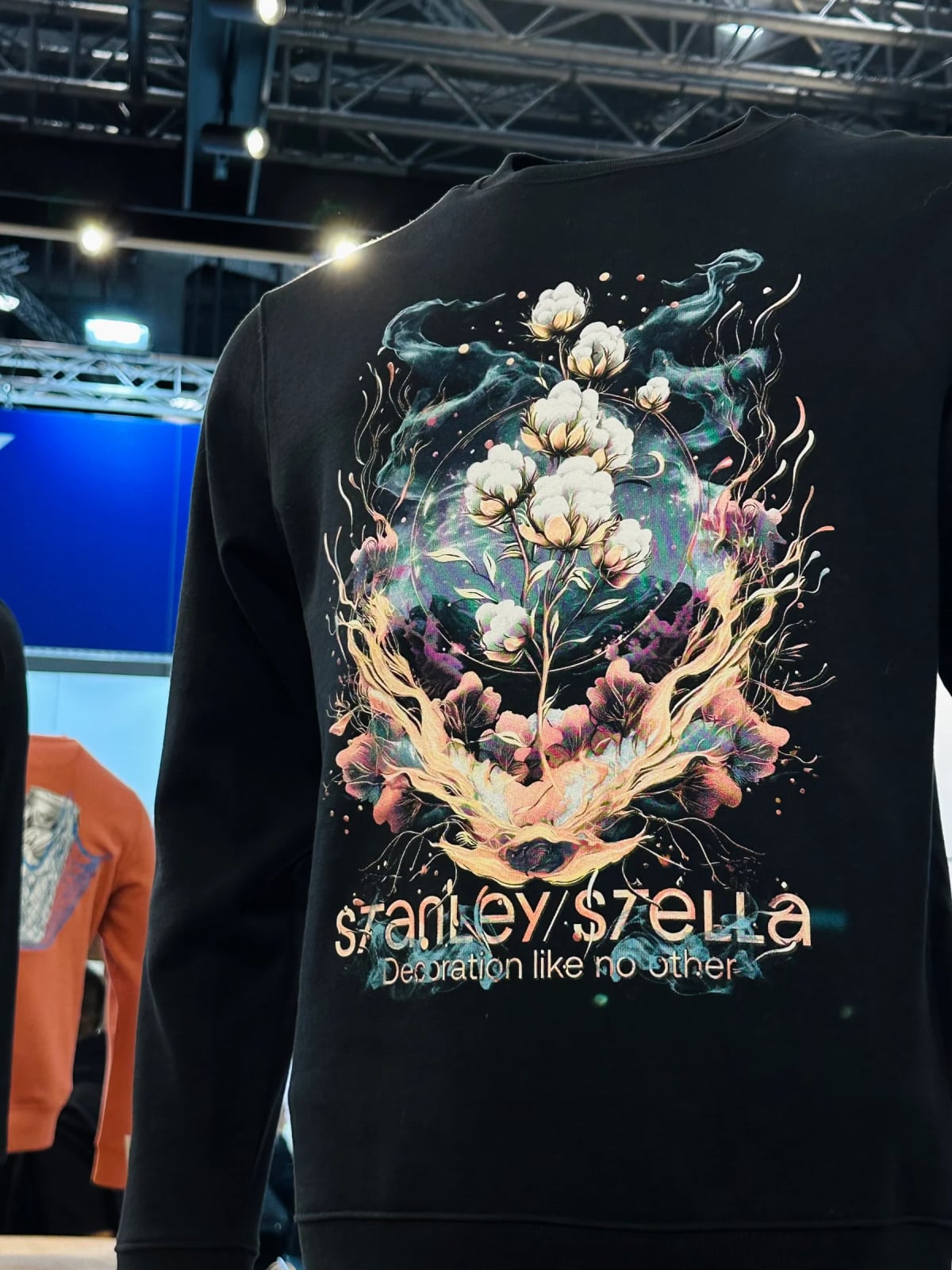 Back view of a black crewneck sweatshirt displayed on a mannequin, featuring a large colorful graphic print of cotton flowers with flame and floral elements, with the text "Stanley/Stella – Decoration like no other" printed below the design.