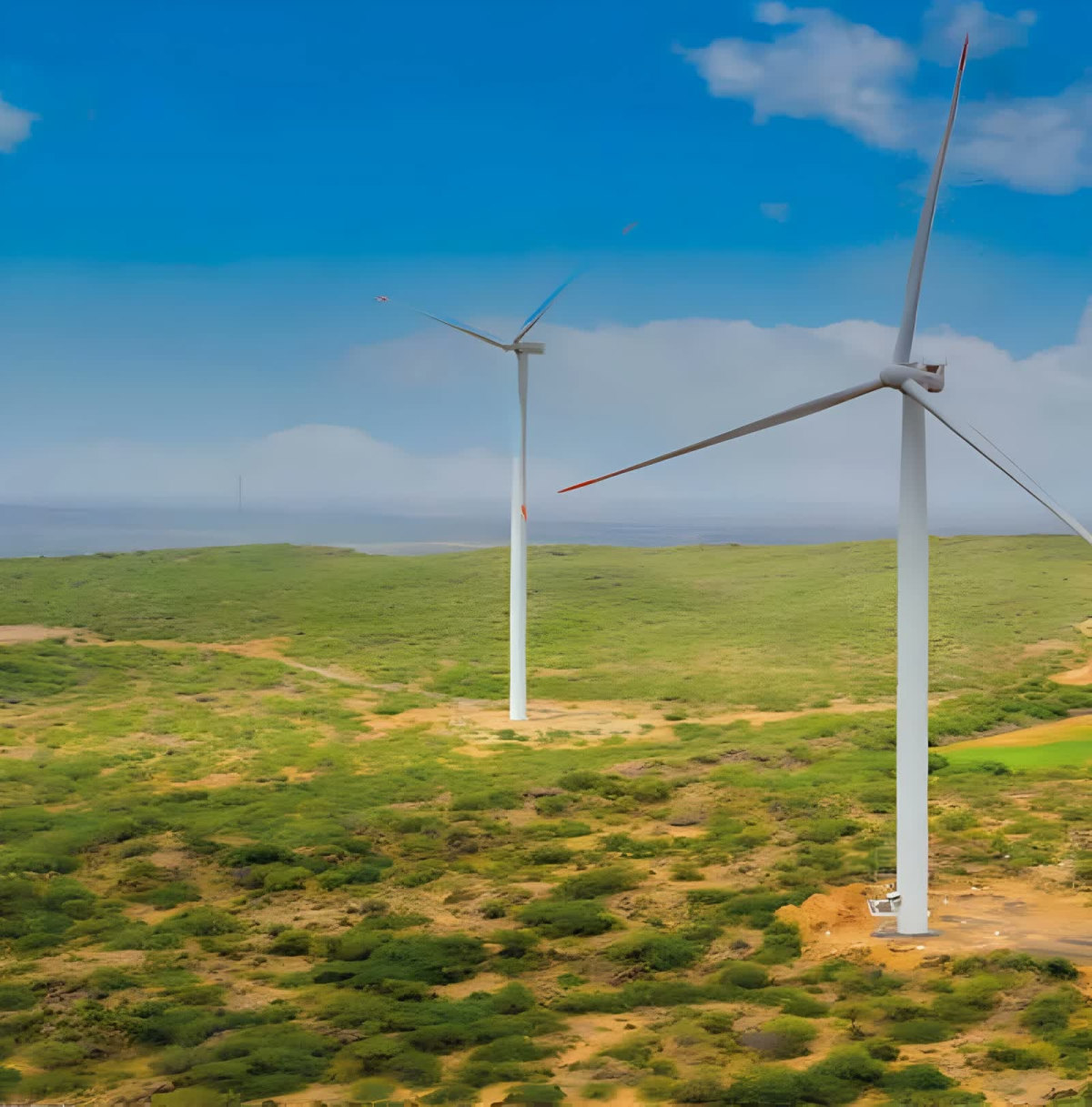 Two large white wind turbines with red-tipped blades stand on a rolling green and sandy landscape under a partly cloudy blue sky, with a second smaller turbine visible in the mid-distance.
