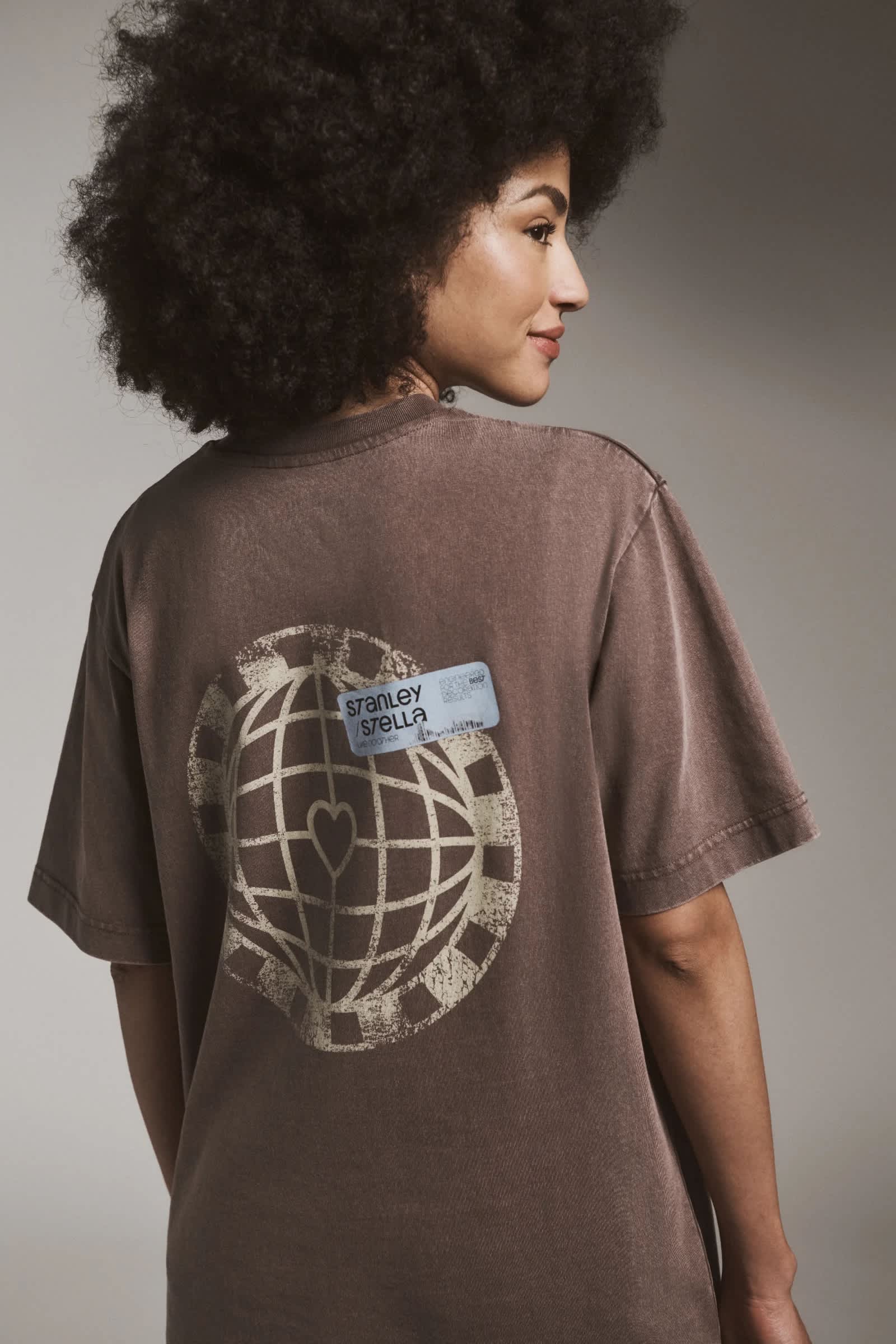 Person wearing a brown Stanley Stella t-shirt with a globe and heart graphic on the back, shown from behind in profile against a neutral gray background.