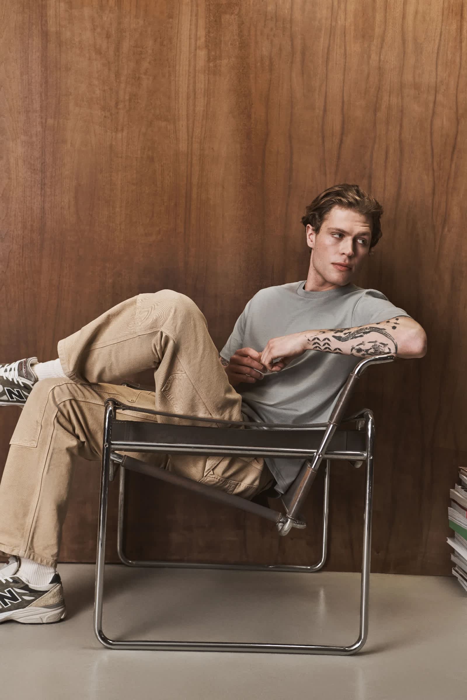 Man sitting on a metal-frame chair against a brown textured backdrop, wearing a gray t-shirt and tan pants.