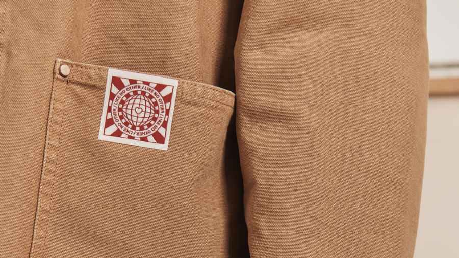 Close-up of a tan canvas jacket showing the front patch pocket, cuff, and a white woven label printed with a red globe motif and the words "Like No Other".