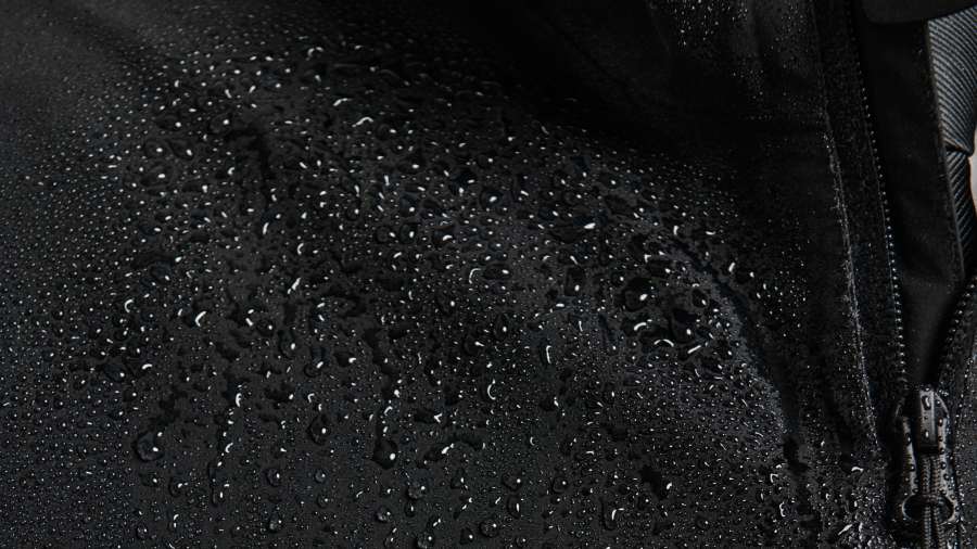 Close-up of a black waterproof jacket covered in water droplets, showing a red circular logo and the white printed text "Sonar Agency" next to a zipper.