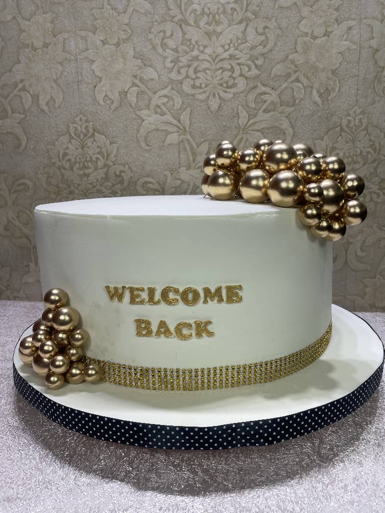 White celebration cake with gold spheres and a welcome back message