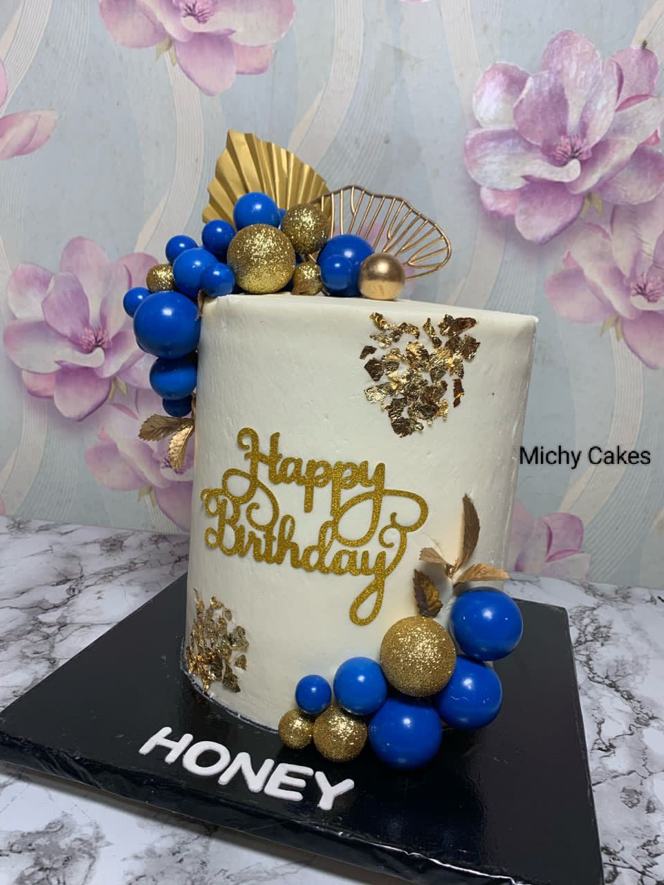 Tall white cake with gold leaf, blue and gold spheres, a gold fan and a happy birthday topper