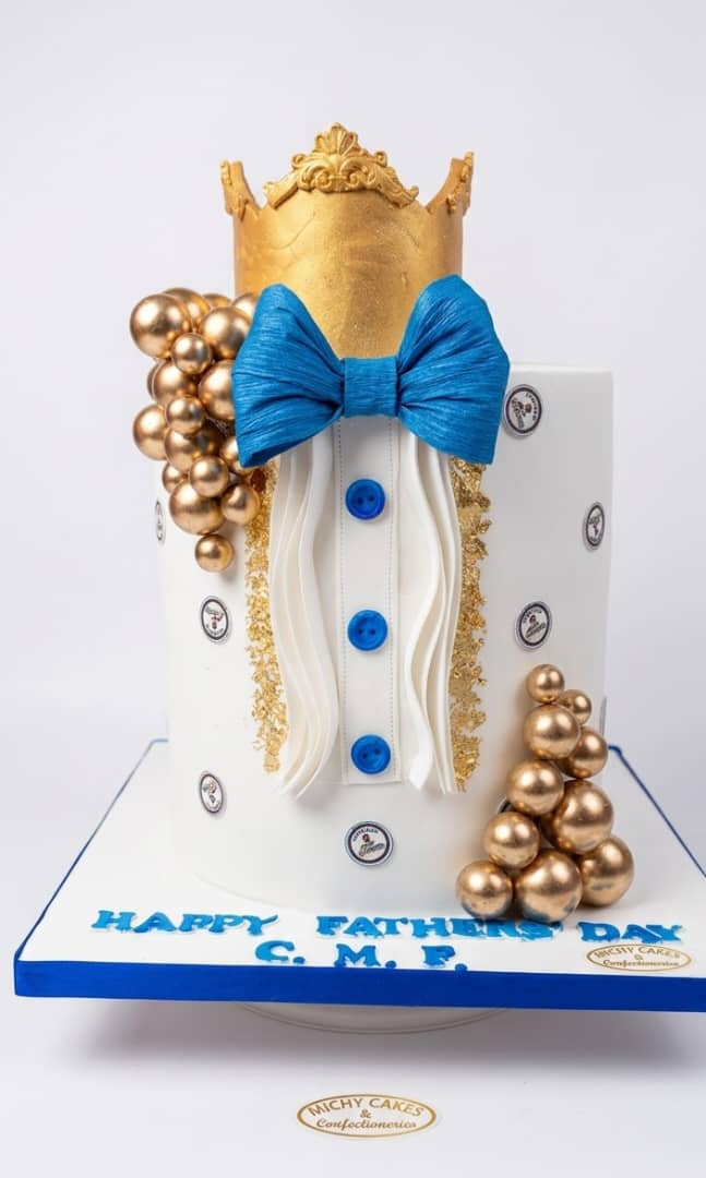 White cake with a gold crown, a blue bow tie, a shirt front with blue buttons and gold spheres