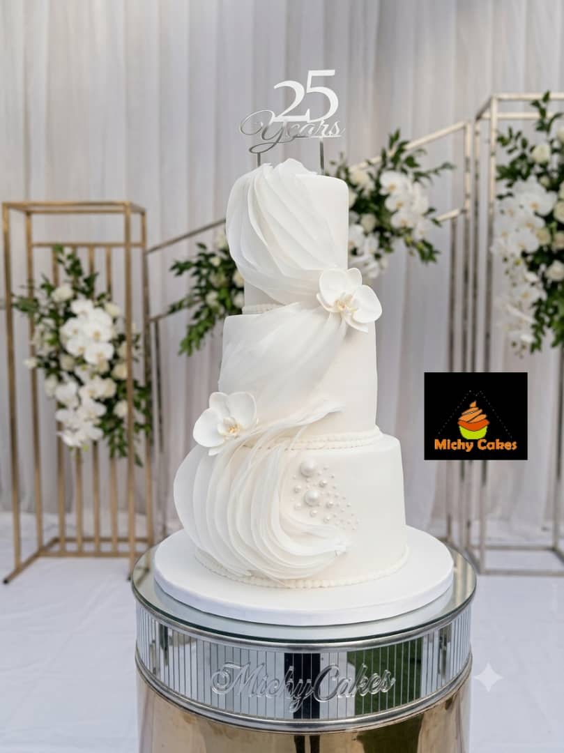 Three-tier white cake with sheer draped ruffles, white orchids, pearls and a silver 25 Years topper