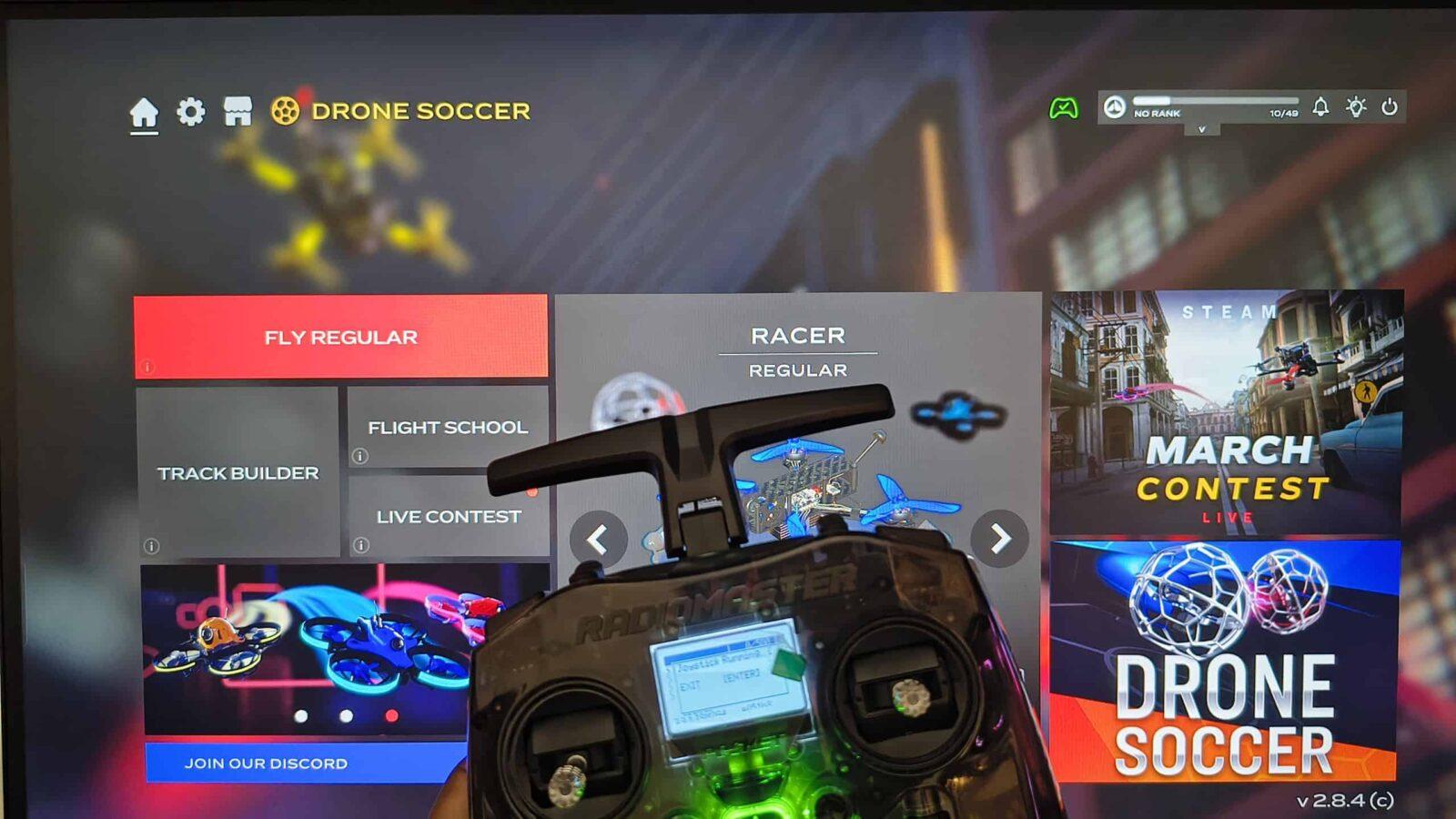A RadioMaster Pocket RC controller held in front of a computer screen displaying the main menu of a "Drone Soccer" simulation game, showing the "Fly Regular" and "Racer" game modes. The simulator is FPV.SkyDive