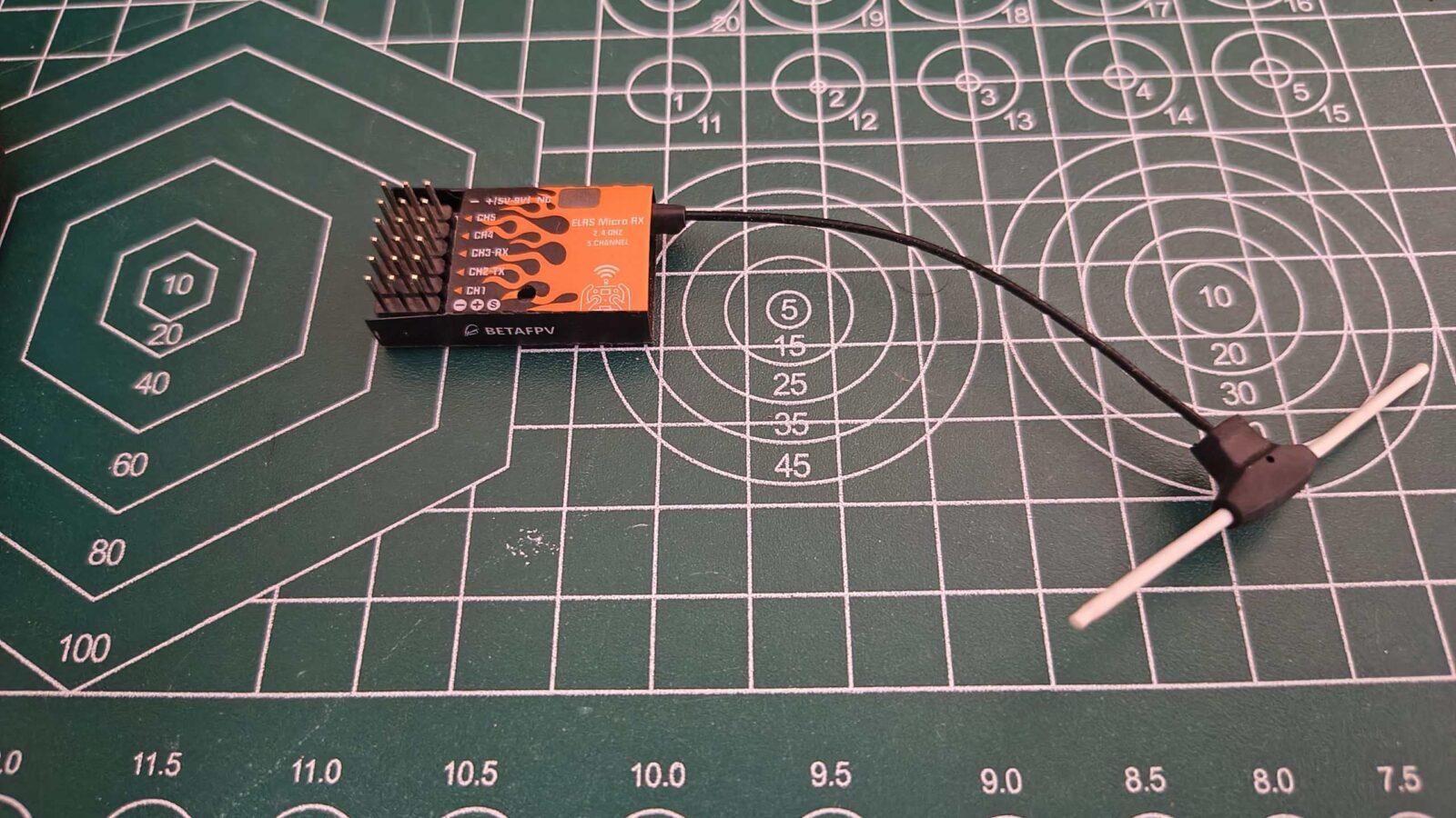 A BETAFPV ELRS Micro RX 5-channel 2.4GHz receiver with an orange and black flame design, connected to a black and white T-style dipole antenna on a green self-healing cutting mat.