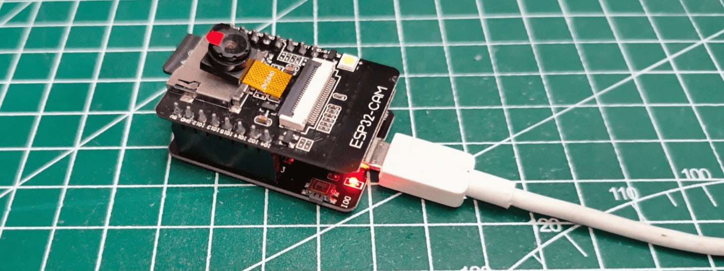 A high-angle close-up shot shows an ESP32-CAM development board connected to a white USB cable. The board, featuring a small camera lens and microSD card slot, sits on a green self-healing cutting mat with white grid lines. A red power LED is illuminated on the board's surface.