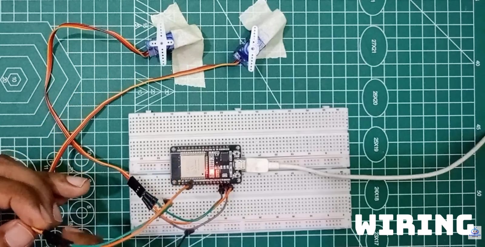 A top-down view of a green cutting mat featuring an ESP32 development board on a white breadboard. Two blue SG90 micro servo motors are secured with tape at the top, with their wiring connected to the ESP32. A white USB cable powers the board, and a hand is visible at the bottom left adjusting a con