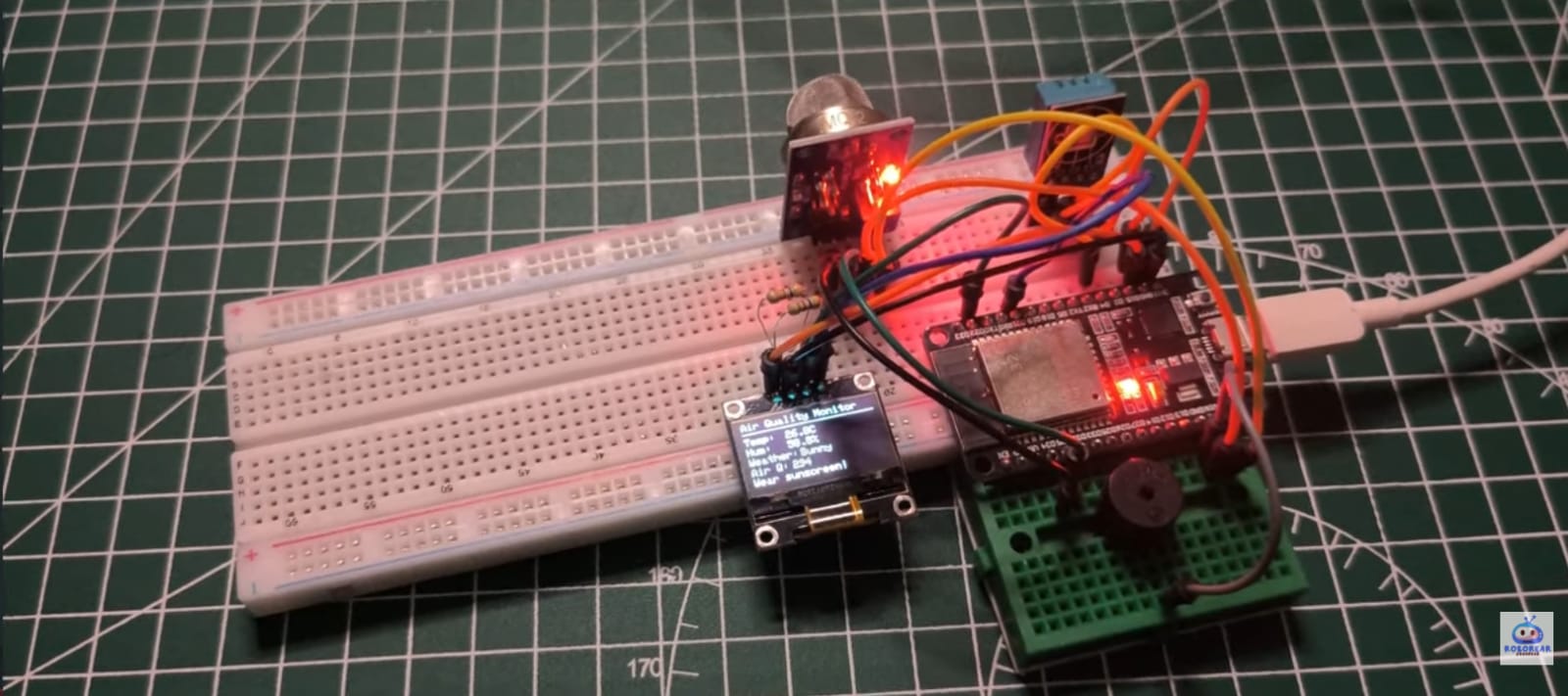 An ESP32 microcontroller mounted on a breadboard over a green cutting mat, functioning as a DIY weather station. The setup includes an MQ gas sensor, a DHT temperature and humidity sensor, and a small OLED screen displaying live data: "Temp: 26.9C", "Hum: 39.8%", "Weather: Sunny", and "Wear sunscree