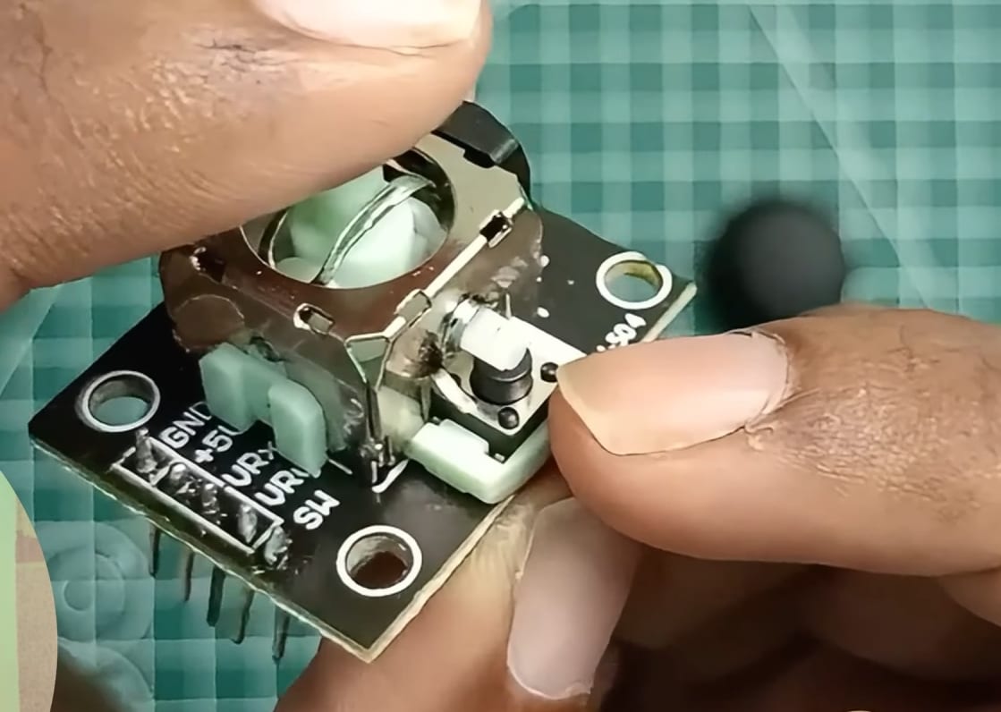 An extreme close-up view of two hands carefully holding a disassembled analog joystick module over a green grid mat. The black thumbstick cap is off to the side, exposing the white mechanical center and the two green side potentiometers. Clear pin labeling for GND, +5V, VRx, VRy, and SW is visible o