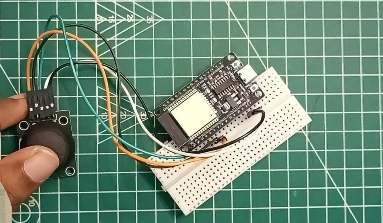A top-down view on a green cutting mat showing a black analog joystick module being held by a thumb, connected via four jumper wires (orange, green, black, white) to an ESP32 development board. The ESP32 is mounted on a small white breadboard, illustrating a clean prototype setup for a Joystick with