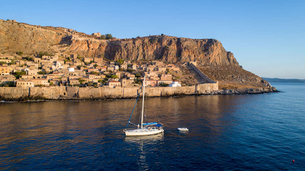 Sailboat sailing near small settlement Monomvasia located on small rocky peninsula next to Peloponnese, Greece. View from the sea at morning, with soft sunlight