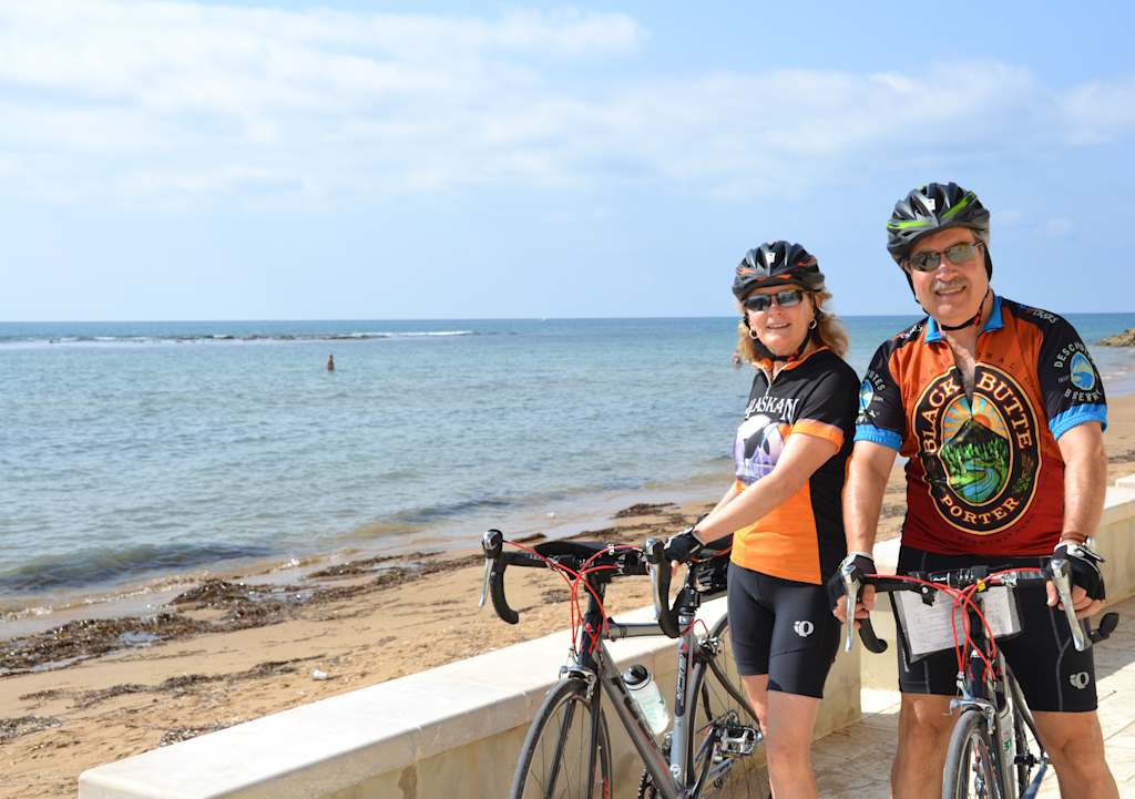 Two cyclists in cycling gear stand on a beach with the ocean visible in the background.