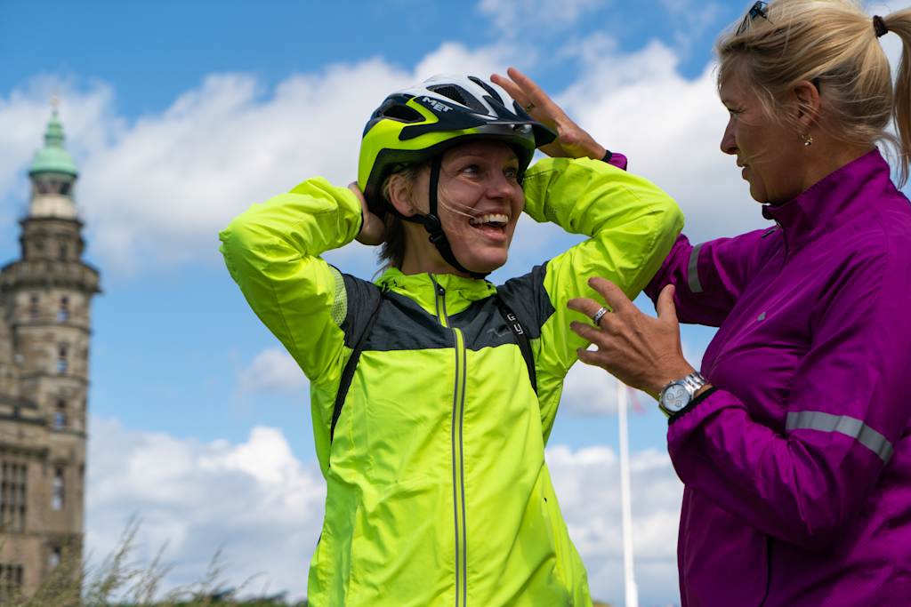 A woman in a bright yellow and black cycling jacket is adjusting her helmet, with a historic building visible in the background.