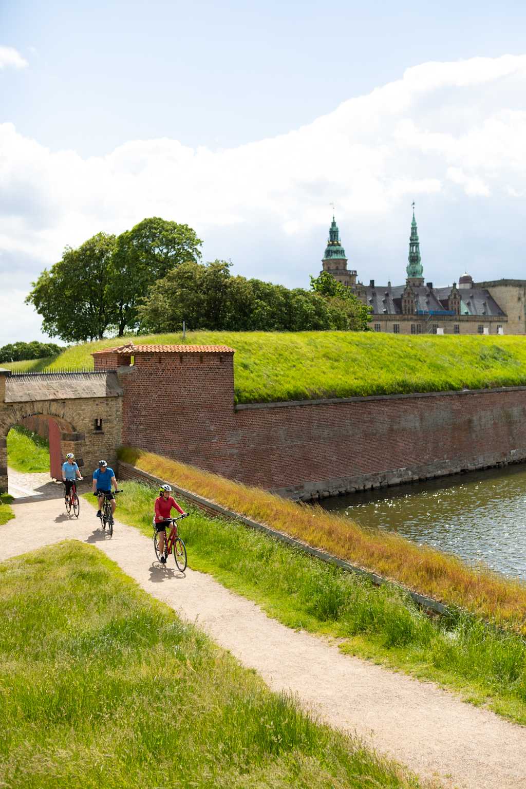 A scenic landscape with a brick wall, a path, and cyclists riding along a grassy area, with a castle-like structure visible in the background.