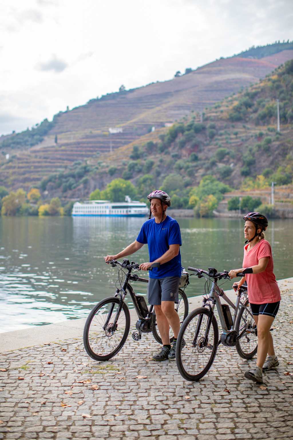 Two people on bicycles stand on a cobblestone path by a body of water, with rolling hills and vineyards visible in the background.