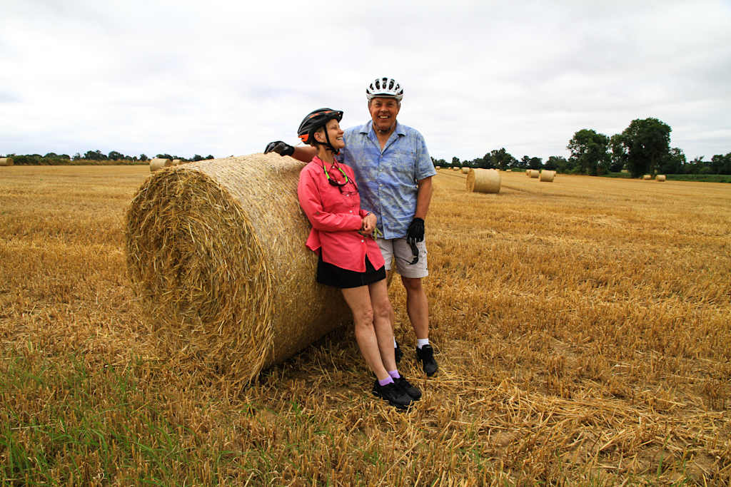 Two people, a man and a woman, standing in a field next to a large hay bale, with a rural landscape in the background.
