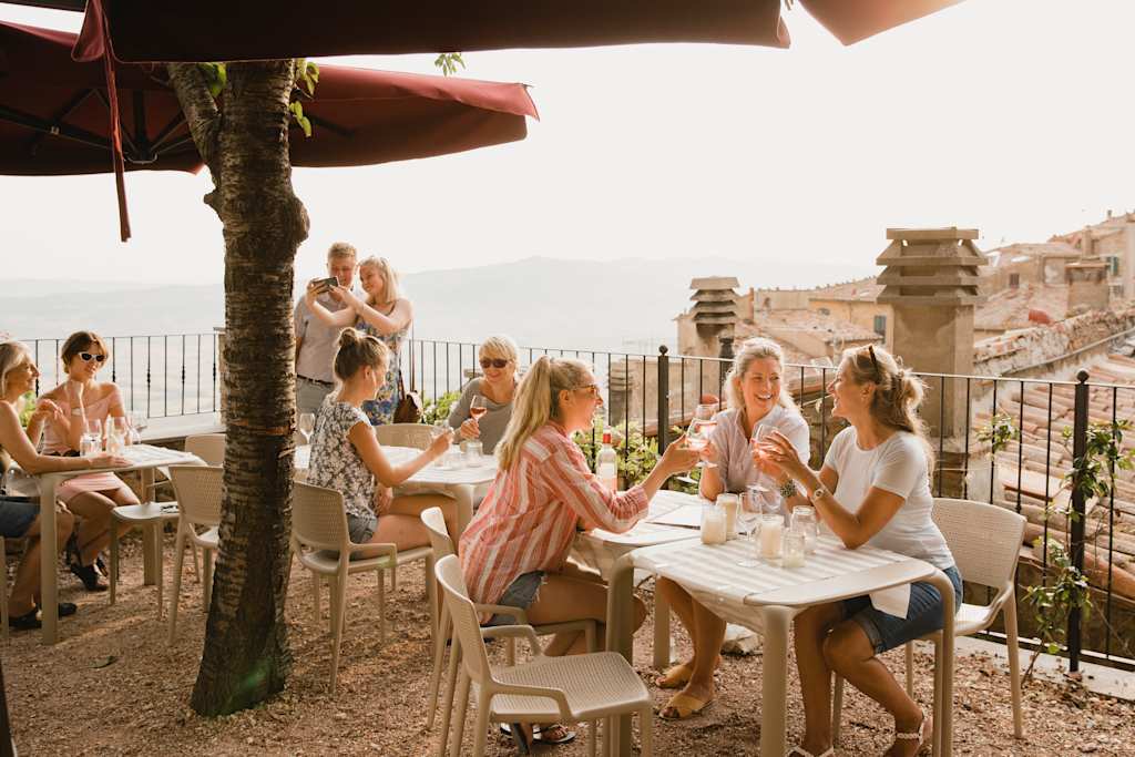 Large group of people sitting and standing on a rooftop terrace of a restaurant in Volterra. Three female friends are making a celebratory toast with their glasses of wine.