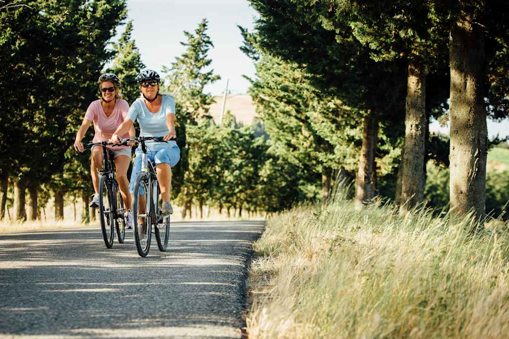 Two mature female friends cycling along a road during a summer holiday in Tuscany. Both wearing cycle helmets and sunglasses as the smile.