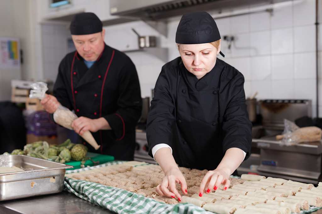 Two chefs in black uniforms and hats are working in a kitchen, one chopping vegetables and the other preparing dough or other ingredients on a counter.