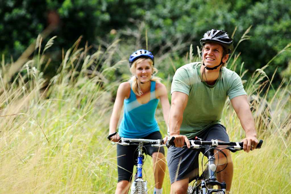 A couple on bicycles riding through a grassy field with lush vegetation in the background.