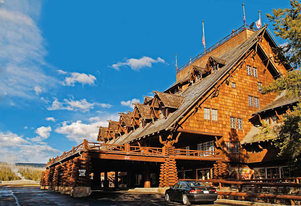A large, rustic wooden lodge with a distinctive peaked roof stands in a scenic outdoor setting, surrounded by trees and a clear blue sky with fluffy white clouds.