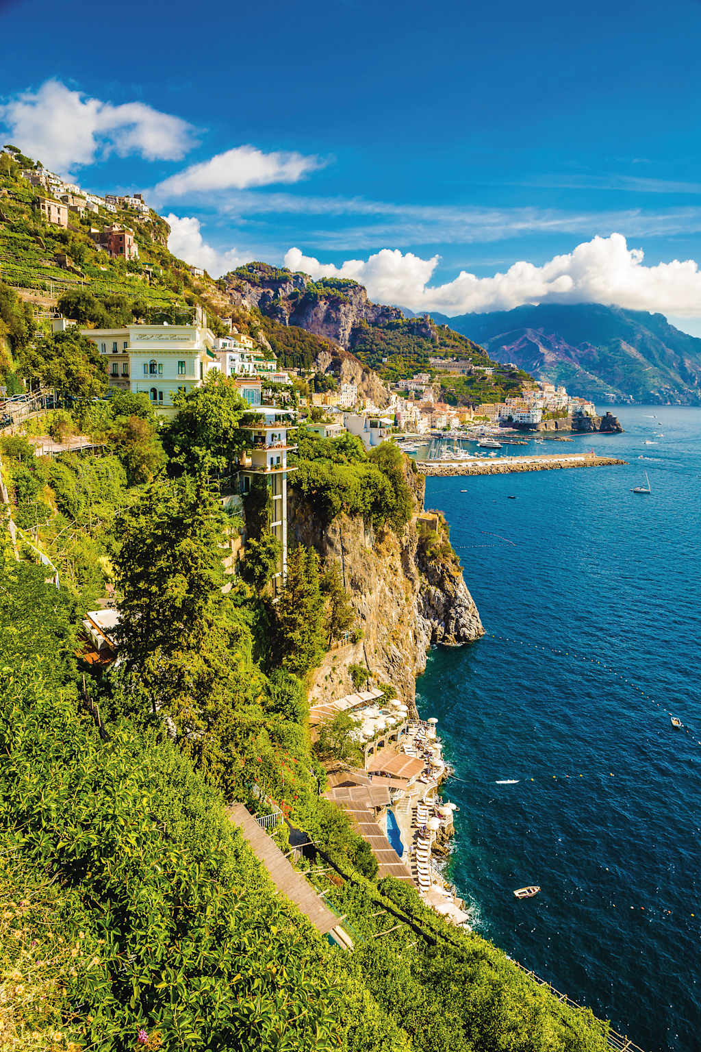 A picturesque coastal town nestled among lush greenery, with colorful buildings cascading down the hillside and a serene blue sea stretching out in the background.