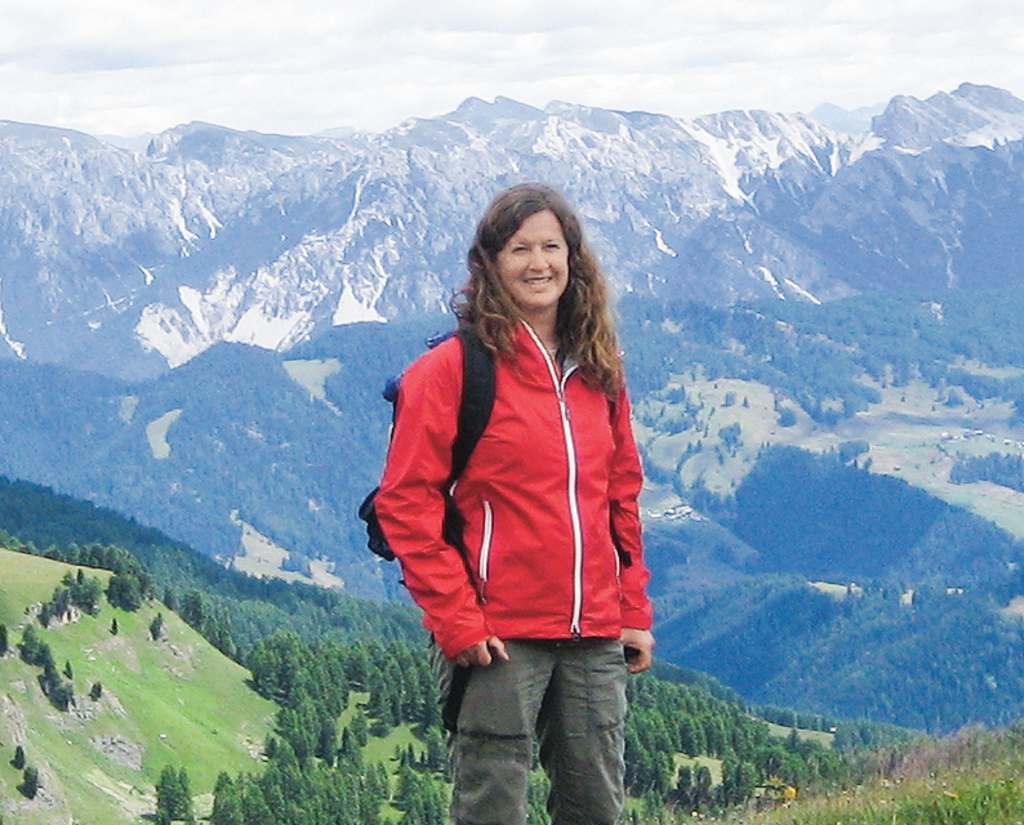 A woman in a red jacket stands in the foreground, with a breathtaking mountain landscape in the background.