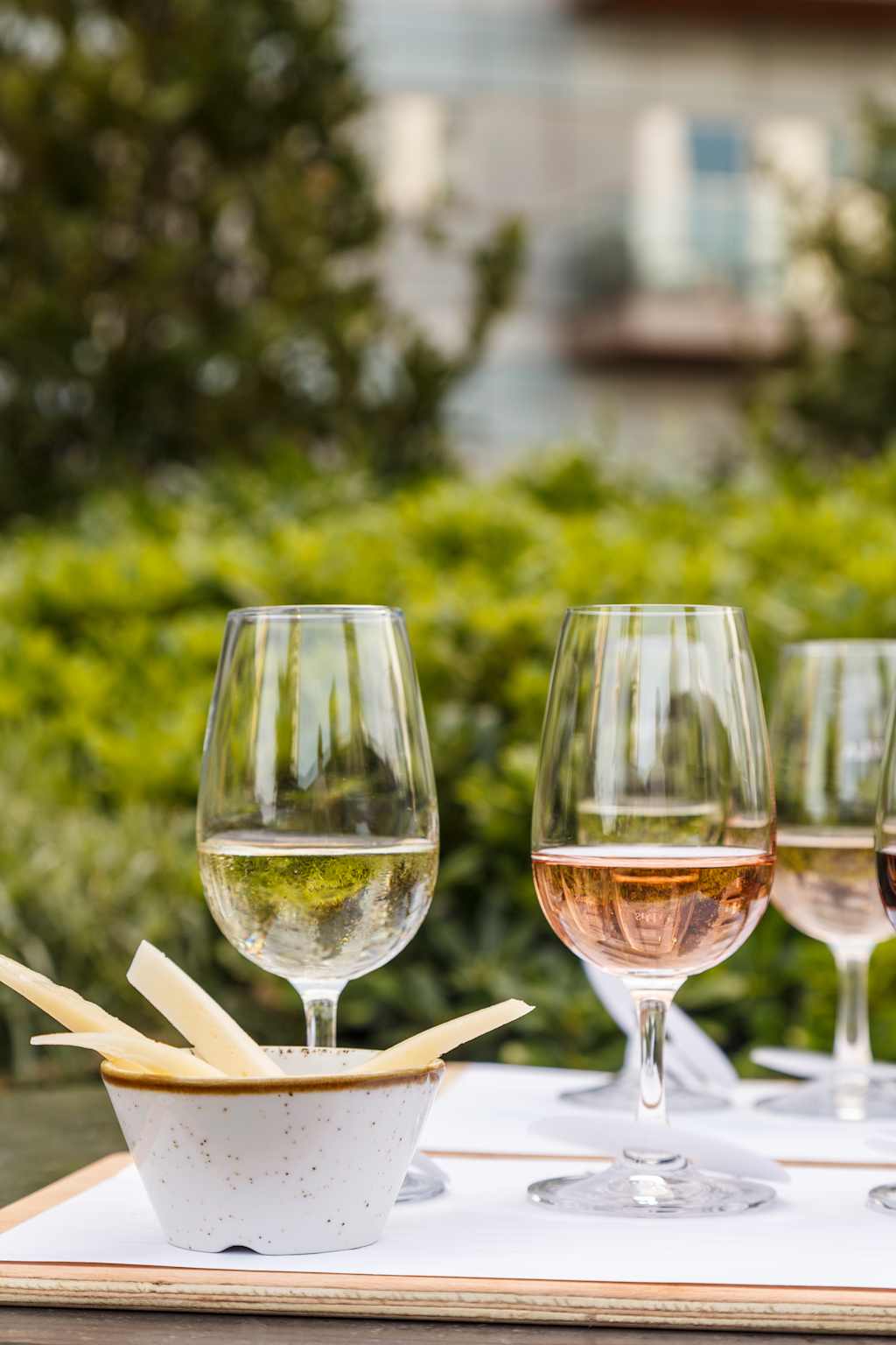 Two wine glasses filled with white and rosé wine, accompanied by a small bowl of breadsticks, set against a blurred background of lush greenery and a building.