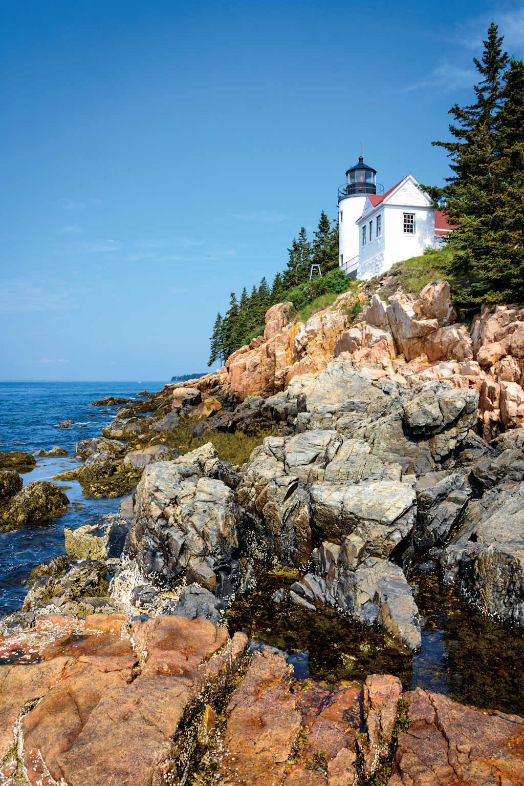 A picturesque lighthouse stands atop a rocky coastal outcrop, surrounded by a rugged shoreline and a vast expanse of the ocean under a clear blue sky.