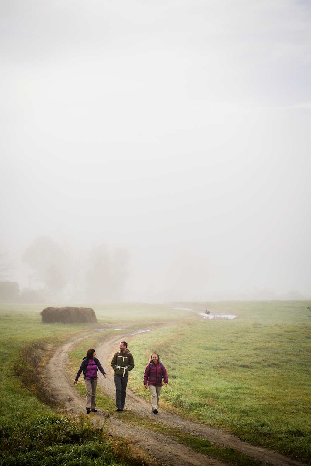 Three people walking on a dirt path through a misty, grassy field with a few bales of hay in the background.