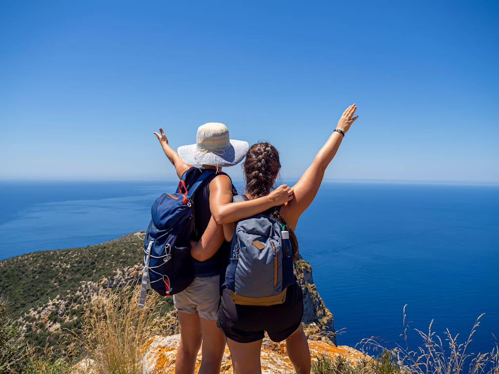 Two hikers with backpacks stand on a cliff overlooking a vast, blue ocean against a clear sky.