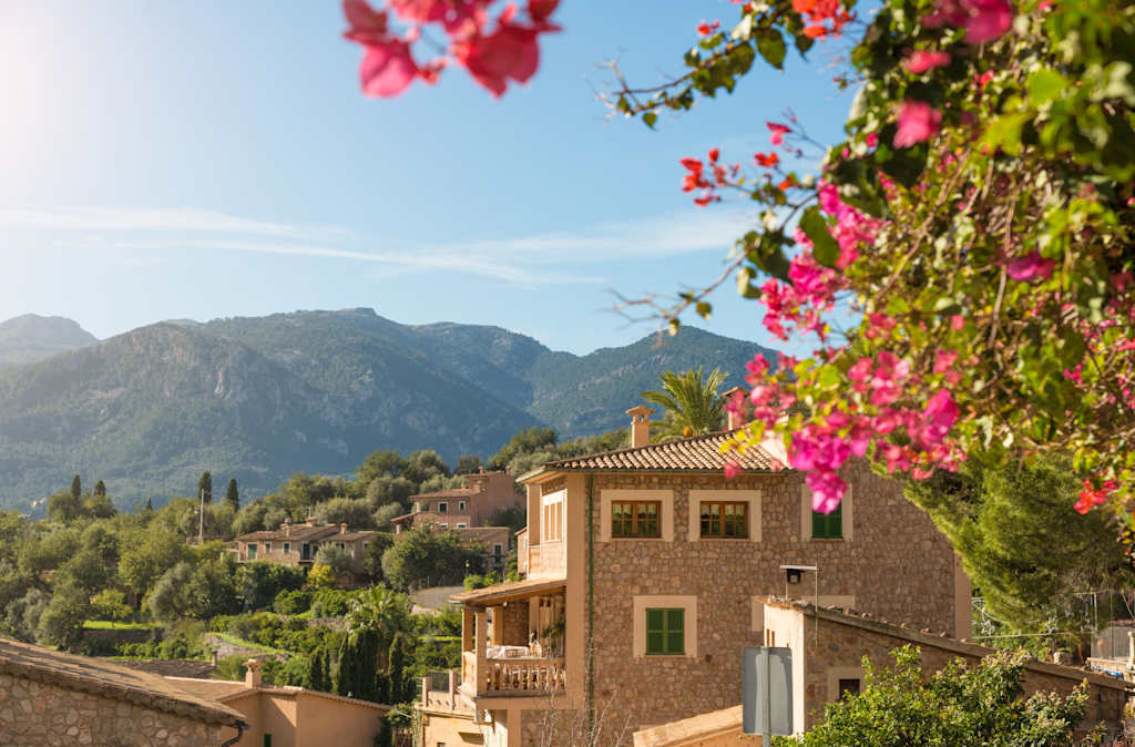 A picturesque Mediterranean-style villa nestled among lush greenery, with vibrant bougainvillea flowers cascading over the balcony, set against a backdrop of majestic mountains.