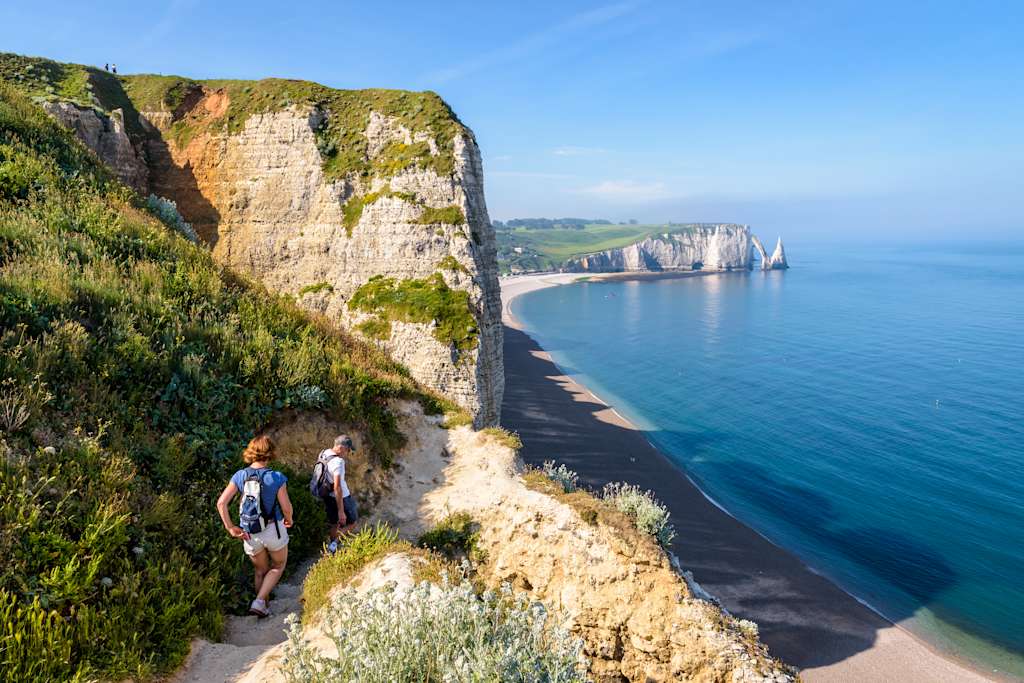 Rugged cliffs overlooking a tranquil blue sea, with two people hiking along a winding path in the foreground.